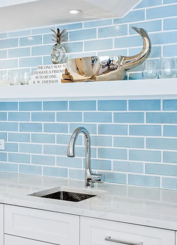 A bar with light blue tiling black splash. The sink has a faucet with a silver head and long neck. There are various cups and vases decorating the shelves and counter top.